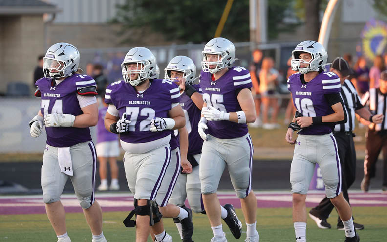 Five people wearing purple and white football uniforms and helmets are walking together on a football field. Description generated by AI