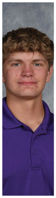 A person with curly hair wearing a purple collared shirt is standing against a gray textured background. Description generated by AI
