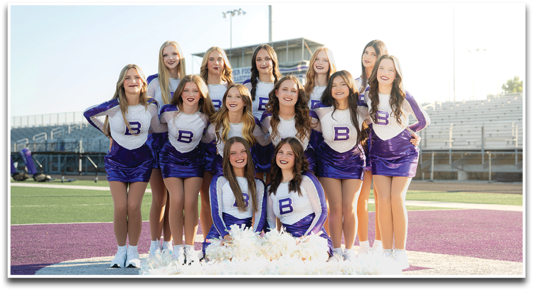 A group of people wearing matching purple and white cheerleading uniforms pose together on a sports field with smiles. Description generated by AI