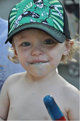 A child wearing a Green Lantern cap is holding a partially eaten blue and red popsicle with some melted popsicle on their face. Description generated by AI