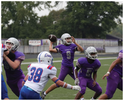 Players in purple and white football uniforms are engaged in a game on a green field with trees in the background. Description generated by AI