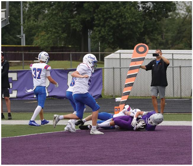 Several people in football uniforms are on a field with some lying on the ground and others standing or running near an orange and black striped marker. Description generated by AI