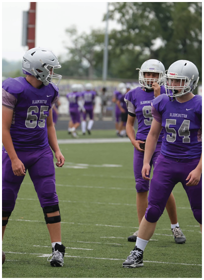 Three people in purple football uniforms and silver helmets stand on a football field with one holding a football. Description generated by AI