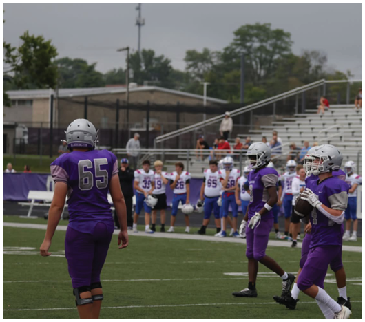 People in purple football uniforms are on a field with others in white and blue uniforms standing on the sidelines in the background. Description generated by AI