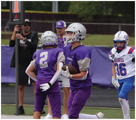 Two people in purple football uniforms are on a field with one holding a football while a person in a white and blue uniform stands nearby and a person in black holds a sign with the number one. Description generated by AI