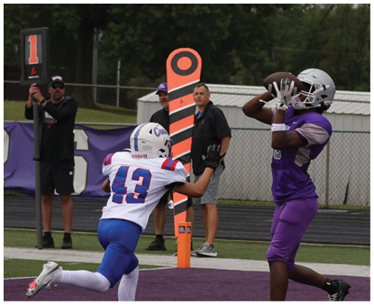 A person in a purple sports uniform and white helmet is catching a football while two people hold up yard markers on a grassy field with trees in the background. Description generated by AI