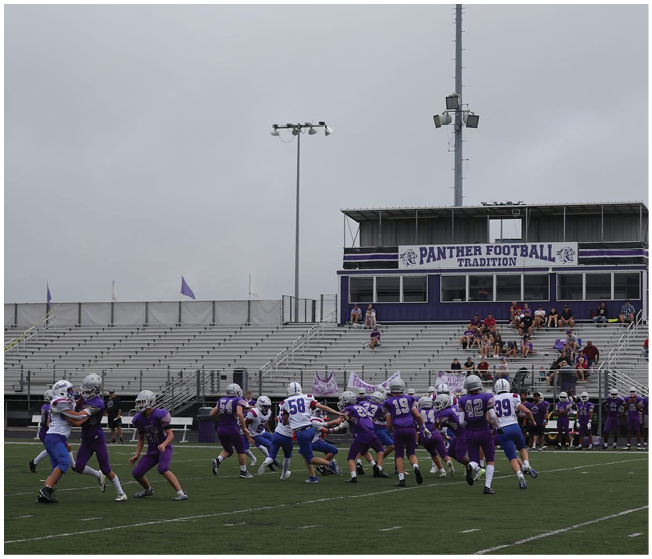 A group of people in football uniforms are playing on a field in front of empty bleachers and a press box with a sign that reads Panther Football Tradition. Description generated by AI