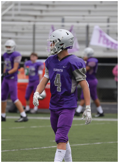 A person wearing a purple football uniform with the number 4 and a silver helmet is standing on a football field with other players in the background. Description generated by AI