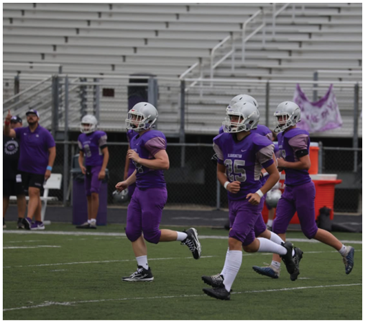 Three people in purple football uniforms and helmets are running on a field with a few others and a coach in the background near bleachers. Description generated by AI