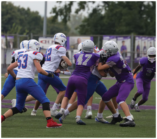 People wearing football uniforms are engaged in a play on a grassy field with some players blocking and others preparing to move. Description generated by AI