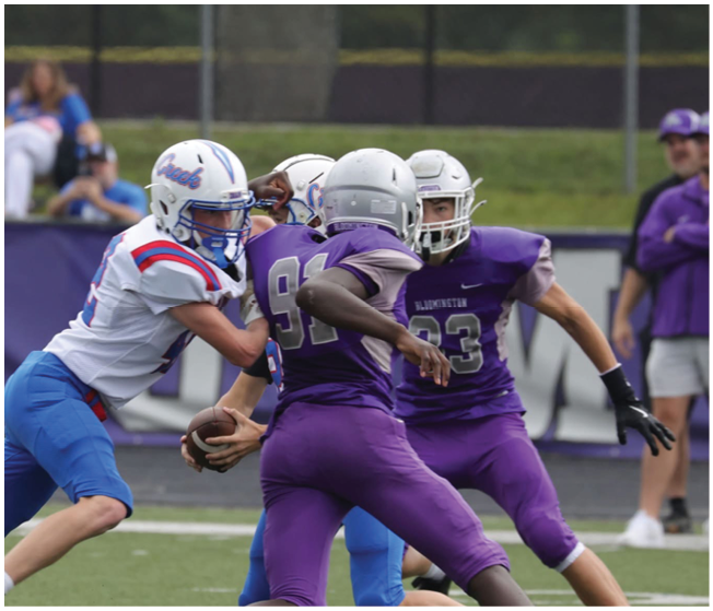 Two groups of people wearing football helmets and uniforms are engaged in a play on a grassy field with a blurred background of spectators and fencing. Description generated by AI