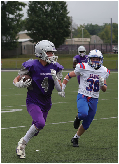 A person in a purple football uniform is running with the ball while being chased by a person in a white and blue football uniform on a grassy field. Description generated by AI