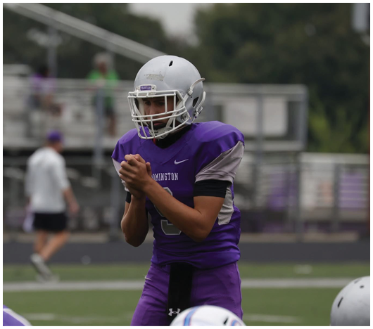 A person wearing a white football helmet and purple jersey is on a field with blurred trees and spectators in the background. Description generated by AI