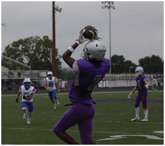 A person wearing a purple sports jersey and white helmet is catching a football outdoors under an overcast sky. Description generated by AI