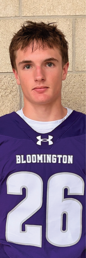 A person with short hair wearing a purple sports jersey with the number 26 and the word Bloomington stands against a stone wall. Description generated by AI