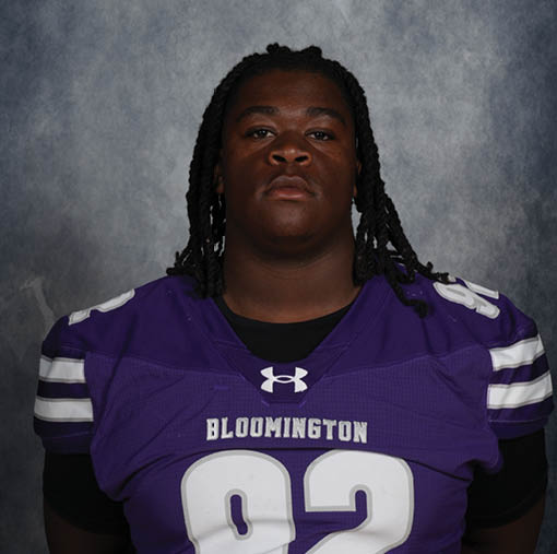 A person with braided hair wearing a purple sports jersey with white stripes and the word Bloomington on the front is posing against a gray textured background. Description generated by AI