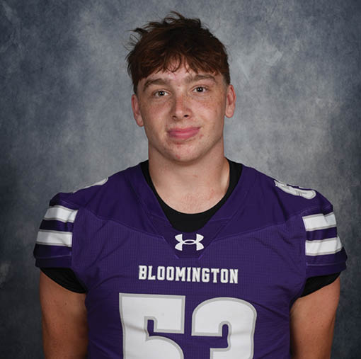 Person with short tousled hair wearing a purple sports jersey with the word Bloomington on it against a gray textured background. Description generated by AI