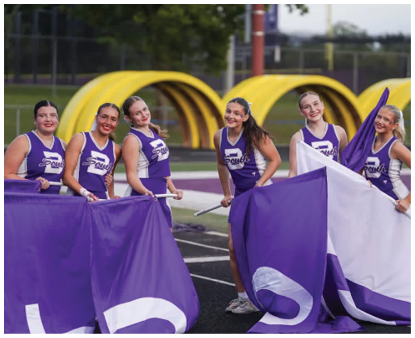 Five adults in matching purple and white uniforms hold large purple flags while standing on a track field with yellow playground equipment in the background. Description generated by AI