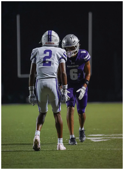 Two people in football uniforms and helmets face each other on a field at night, one wearing a white jersey with the number 2 and the other in a purple jersey with the number 6. Description generated by AI