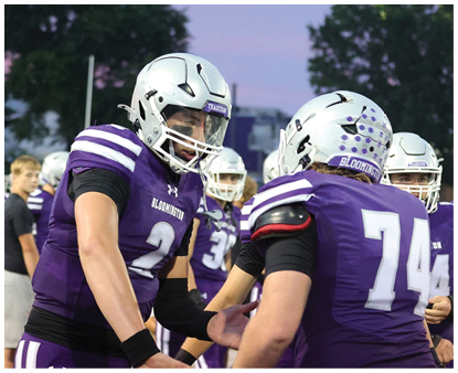 Two people wearing purple football uniforms and silver helmets are interacting on a field with others in the background during a game or practice. Description generated by AI