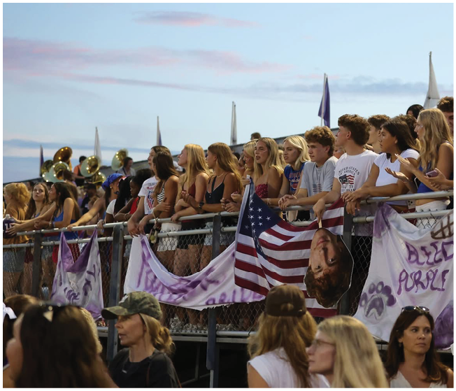 A group of people standing behind a fence decorated with banners and an American flag, watching an event with a band playing in the background during sunset. Description generated by AI