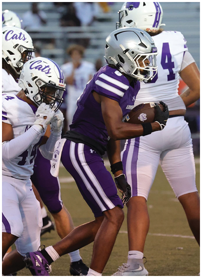 A person in a purple football uniform and silver helmet is running with a football while being pursued by players in white uniforms with “Cats" on their helmets on a football field. Description generated by AI