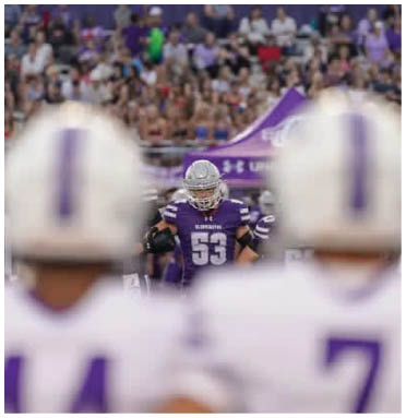 People are sitting and standing on bleachers near a purple and white building with windows, likely at an outdoor event or sports game. Description generated by AI