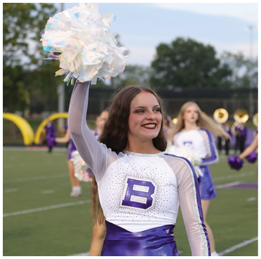A person in a purple and white cheerleading uniform is smiling and holding a white pom-pom up on a sports field with other similarly dressed people and musicians in the background. Description generated by AI