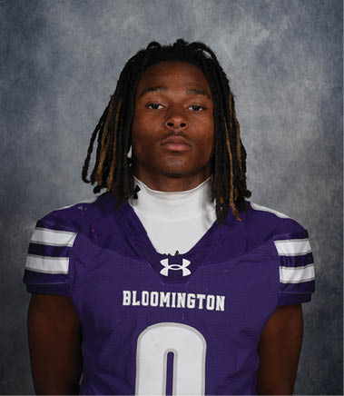 Person with shoulder-length dreadlocks wearing a purple Bloomington sports jersey with the number zero stands against a gray textured background. Description generated by AI