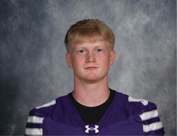 A person with short light hair wearing a purple sports jersey with white stripes stands against a gray mottled background. Description generated by AI