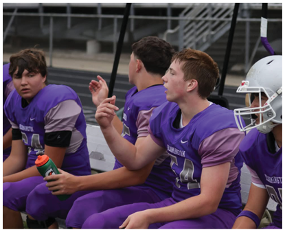 People wearing purple sports uniforms sit on a bench with one holding a green and orange water bottle and a silver helmet on the ground. Description generated by AI