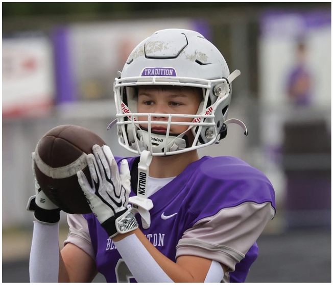 A person wearing a white football helmet and purple jersey is holding a football in a ready-to-throw position. Description generated by AI