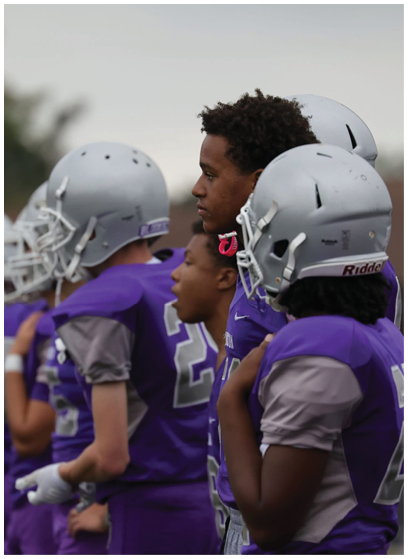 A group of people wearing purple football uniforms and silver helmets stand closely together, appearing focused and attentive. Description generated by AI