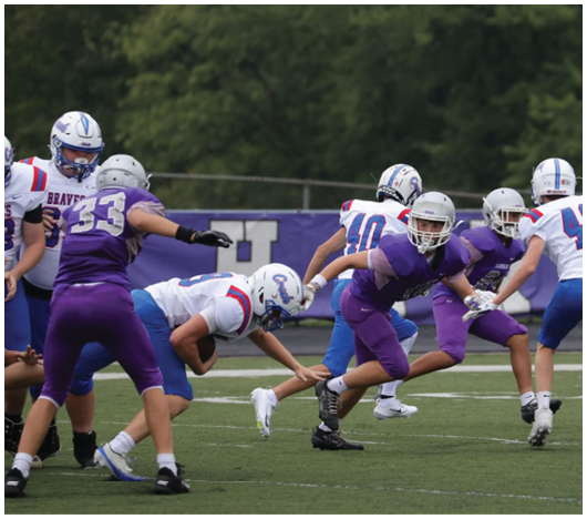 People in football uniforms are actively playing on a field with one person in white and blue being tackled by several people in purple. Description generated by AI