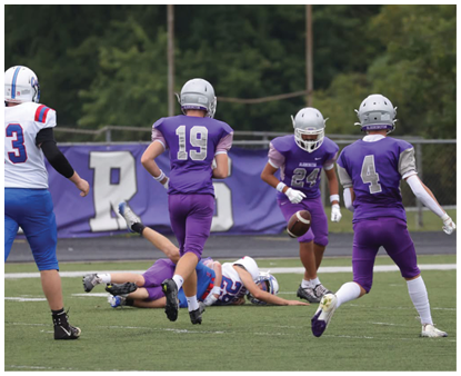 Five people in football uniforms are on a field with one person lying on the ground holding a football while others stand or run nearby. Description generated by AI