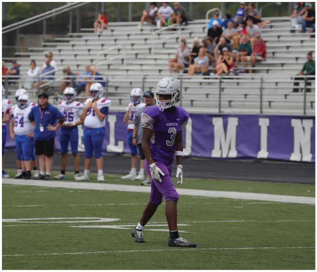 A person in a purple football uniform stands on a field near a group of people in white and blue uniforms with a mostly empty bleacher background. Description generated by AI