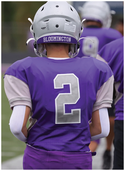 A person wearing a purple football jersey with the number 2 and a white helmet labeled Bloomington stands on a field with other players in the background. Description generated by AI
