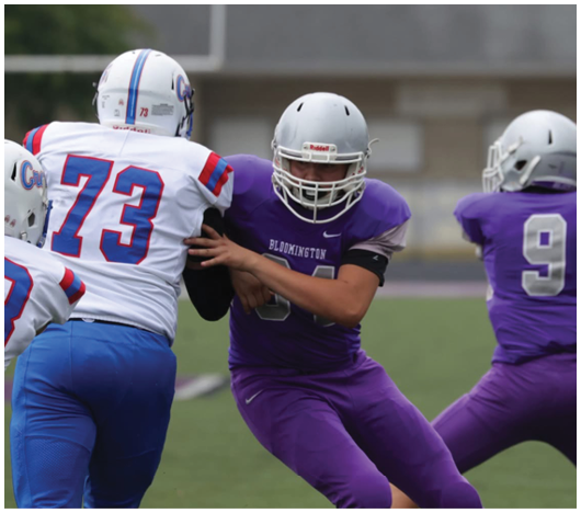 Two people in football uniforms are engaged in a play on a field with one person in purple attempting to block or tackle another person in white and blue. Description generated by AI