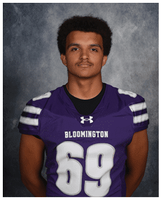 A person with short curly hair wearing a purple Bloomington football jersey with the number 69 stands against a gray textured background. Description generated by AI