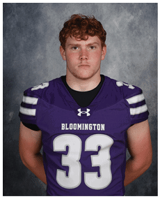 A person with curly hair wearing a purple Bloomington football jersey with the number 33 stands against a gray textured background. Description generated by AI