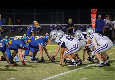 Two opposing American football teams in blue and white uniforms are lined up facing each other on a field under stadium lights, preparing for a play. Description generated by AI