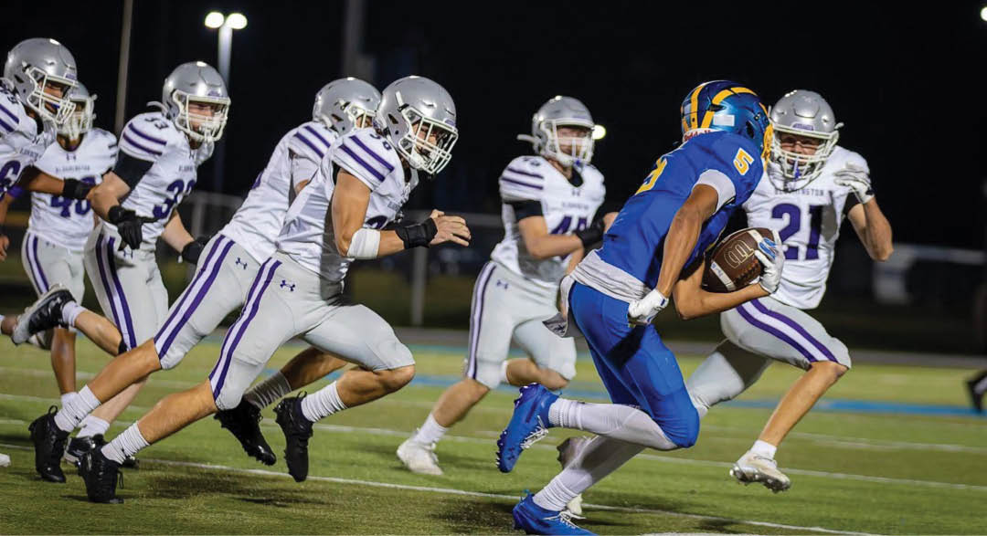 A group of people in white and purple football uniforms are chasing a person in a blue and yellow football uniform who is running with the ball on a field at night. Description generated by AI
