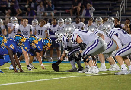 Two opposing American football teams in blue and white uniforms are lined up at the line of scrimmage on a field with spectators in the background. Description generated by AI