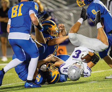 A group of people in blue and yellow football uniforms tackle a person in a white and purple football uniform who is falling upside down on a grassy field. Description generated by AI