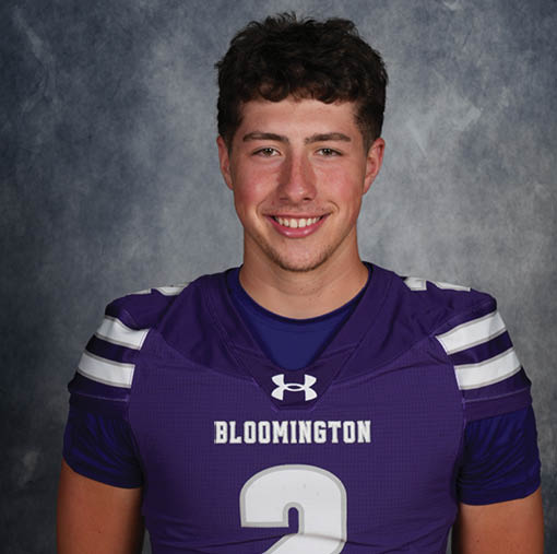A person with short curly hair is smiling and wearing a purple sports jersey with the word Bloomington on it against a gray textured background. Description generated by AI