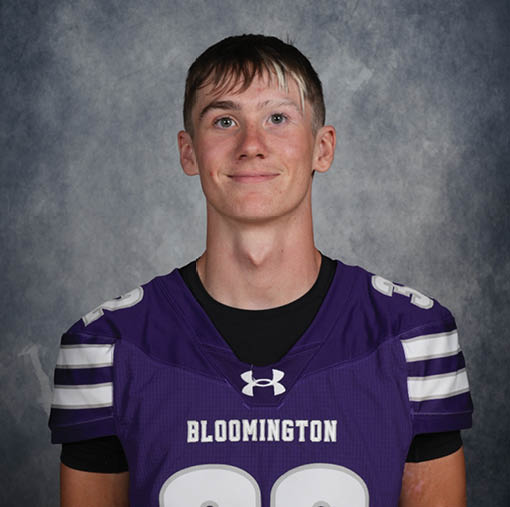 A person with short hair wearing a purple sports jersey with the word Bloomington and the number 22 stands against a gray textured background. Description generated by AI