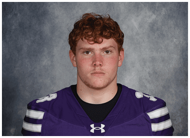 A person with short hair wearing a purple sports jersey with white stripes on the shoulders stands against a gray mottled background. Description generated by AI