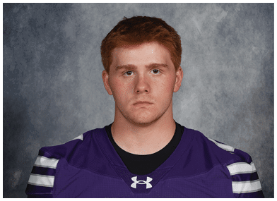 Person with curly hair wearing a purple sports jersey with white stripes and the word Bloomington on the front against a gray mottled background. Description generated by AI