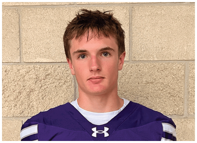A person wearing a purple Bloomington sports jersey with white stripes on the sleeves stands against a beige brick wall. Description generated by AI
