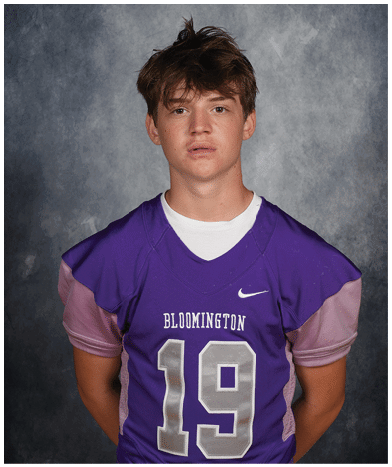 A person with tousled hair wearing a purple Bloomington sports jersey with the number 19 stands against a gray textured background. Description generated by AI